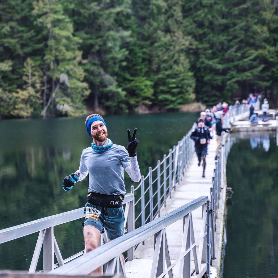 Bastien running across a bridge over a lake during a trail race, smiling and making a peace sign